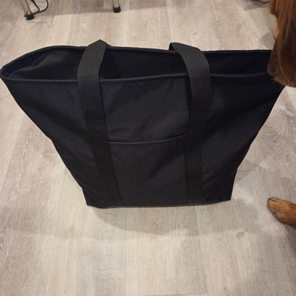 A large black fabric tote bag with two shoulder straps, standing on a light wood floor. A dog's paw is visible at the right edge of the frame.